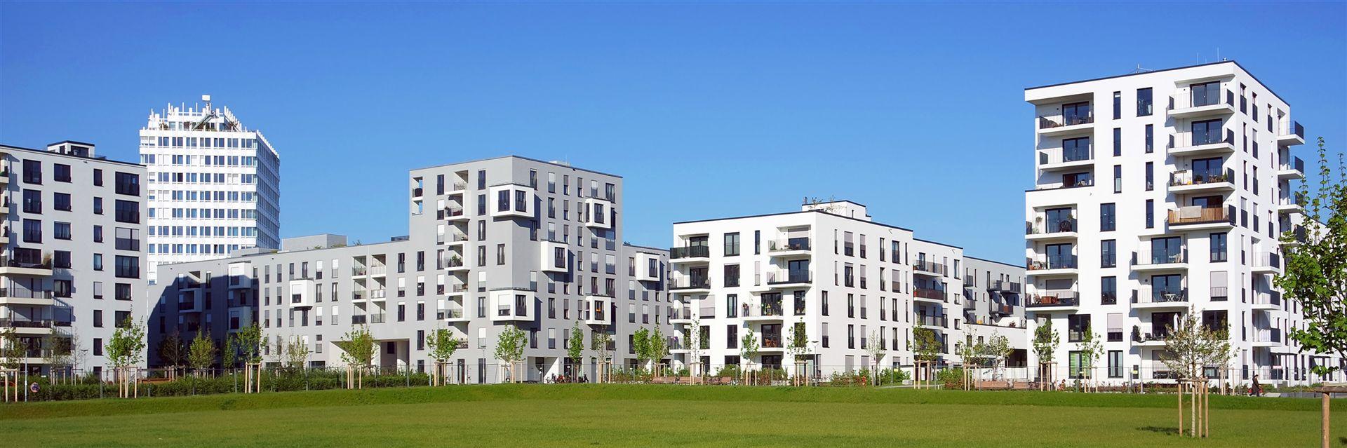 Das Bild zeigt eine moderne Wohnsiedlung mit mehrstöckigen, weißen Wohngebäuden, Balkonen und großen Fenstern vor blauem Himmel und Grünfläche.