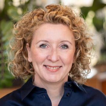 Das Bild zeigt eine Frau mit blonden, lockigen Haaren und dunkler Bluse, im Hintergrund sind Pflanzen sichtbar.