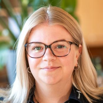 Das Bild zeigt eine Frau mit langen, blonden Haaren und Brille, im Hintergrund sind Pflanzen zu sehen.