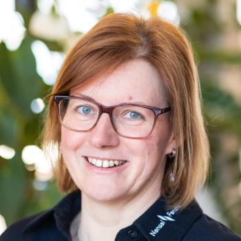 Das Bild zeigt eine Frau mit Brille und rötlich-blonden Haaren, im Hintergrund befinden sich Pflanzen.