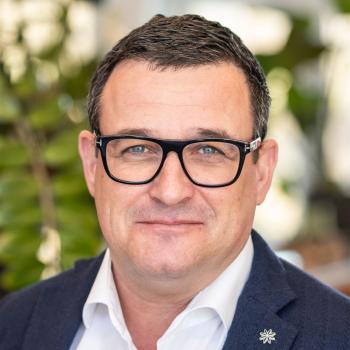 Das Bild zeigt einen Mann mit dunklem Haar und Brille in Businesskleidung, im Hintergrund sind Pflanzen zu sehen.