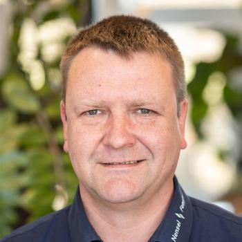 Das Bild zeigt einen Mann mit kurzem Haar in Büro-Umgebung, im Hintergrund sind Pflanzen sichtbar.