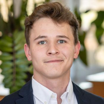 Das Bild zeigt einen jungen Mann mit kurzem, braunem Haar in Businesskleidung, im Hintergrund sind Pflanzen sichtbar.