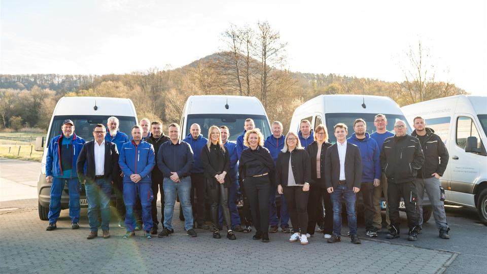 Das Bild zeigt das gesamte Team von Nensel Fensterbau bestehend aus etwa 24 Personen. Die Gruppe steht in zwei Reihen vor mehreren weißen Firmenfahrzeugen auf einem Betriebshof.