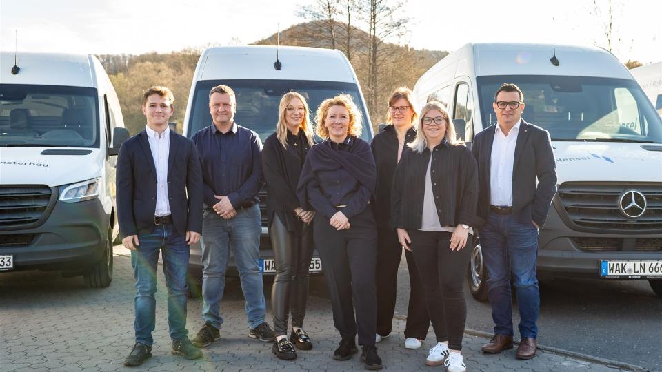 Das Bild zeigt das Büro-Team von Nensel Fensterbau bestehend aus acht Personen, die in Business- und Freizeitkleidung vor den weißen Firmenfahrzeugen stehen. 