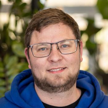 Das Bild zeigt einen Mann mit Brille, kurzem Bart und blauem Kapuzenpullover, der freundlich in die Kamera lächelt.