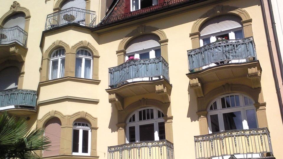 Das Bild zeigt einen Ausschnitt einer Altbaufassade mit verzierten Rundbogenfenstern, Balkonen mit schmiedeeisernen Geländern und geschlossenen Rollläden.