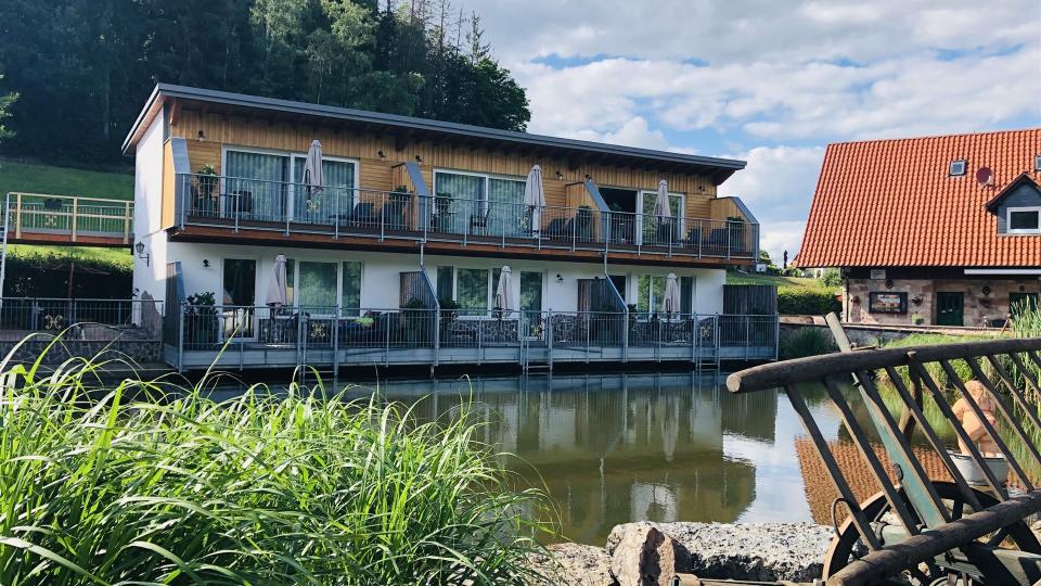 Das Bild zeigt ein modernes Gästehaus mit Holzfassade und Balkonen, direkt an einem kleinen Teich gelegen, umgeben von Natur.