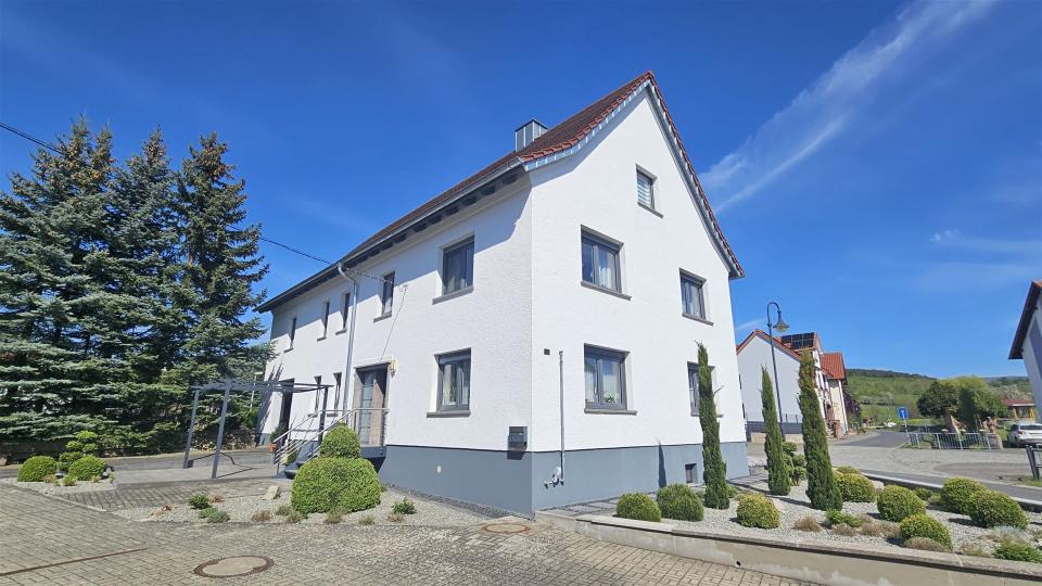Das Bild zeigt ein frisch renoviertes Einfamilienhaus mit weißer Putzfassade, grauem Sockel, anthrazitfarbenen Fenstern und gepflegtem Vorgarten unter blauem Himmel.