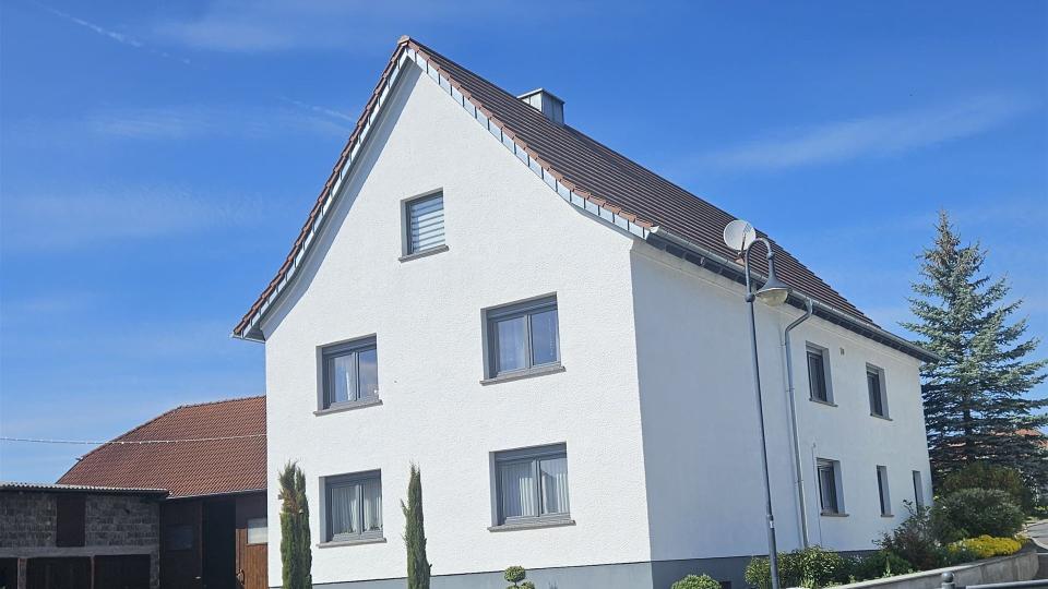 Das Bild zeigt ein frisch renoviertes Einfamilienhaus mit weißer Putzfassade, grauem Sockel, anthrazitfarbenen Fenstern und gepflegtem Vorgarten unter blauem Himmel.