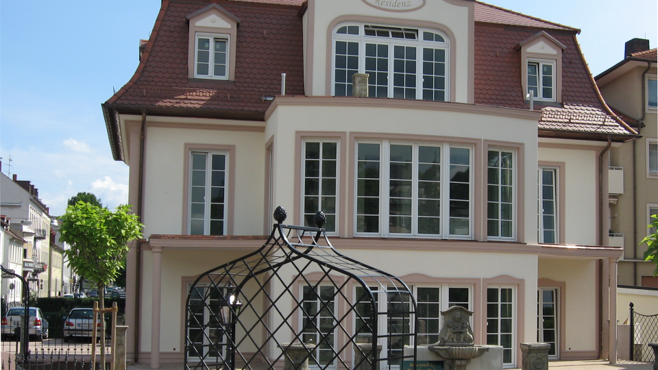 Das Bild zeigt eine elegante Stadtvilla mit Sprossenfenstern, rotem Mansarddach und gepflegtem Vorgarten samt schmiedeeisernem Pavillon.