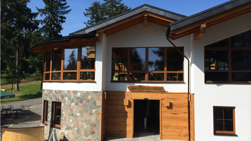 Das Bild zeigt eine moderne Berghütte mit großflächigen Kunststofffenstern in Holzoptik, Naturstein-Sockel und gemütlicher Terrasse mit Metallstühlen, umgeben von Bäumen.