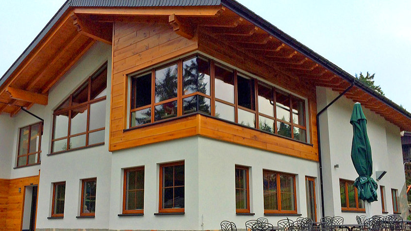 Das Bild zeigt die Märchenwiesenhütte – ein alpines Gebäude mit Naturstein- und Holzfassade, großen Kunststofffenstern in Holzoptik und Satteldach, gelegen in waldreicher Umgebung.