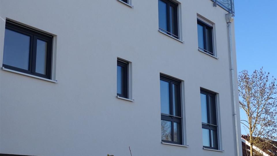 Das Bild zeigt eine moderne, weiß verputzte Hausfassade mit dunklen Fenstern und einem Glasgeländer auf dem Dach, bei klarem blauem Himmel.