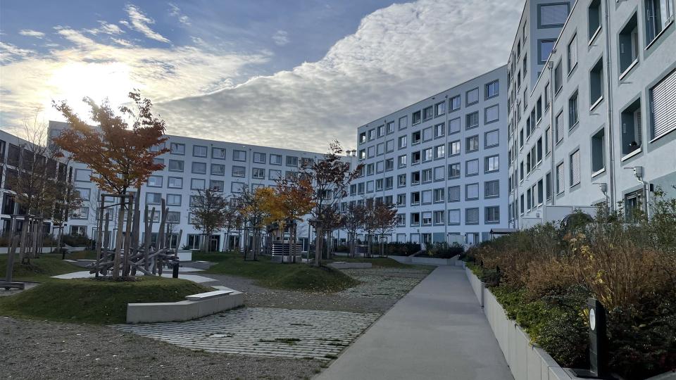 Das Bild zeigt eine moderne Wohnanlage mit weißer Fassade, großem Innenhof, Bäumen in Herbstfärbung und einem Spielplatz unter teils bewölktem Himmel.