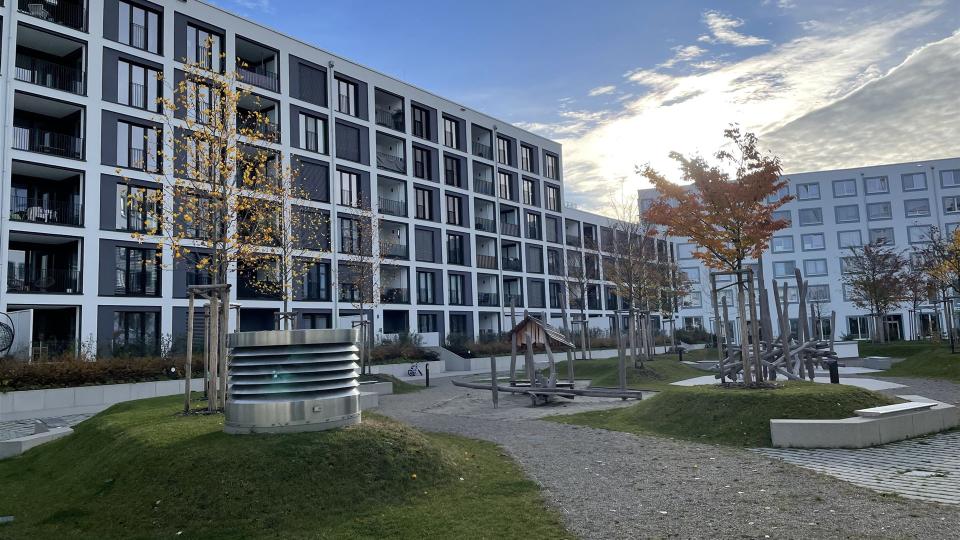Das Bild zeigt eine moderne Wohnanlage mit dunklen Fensterläden, Balkonen und einem begrünten Innenhof mit Spielplatz und Gehwegen bei herbstlichem Wetter.