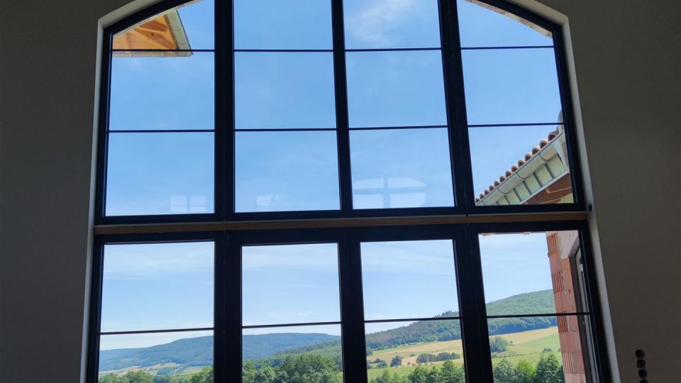 Das Bild zeigt eine große, bogenförmige Fensterfront mit Blick auf eine grüne Landschaft und Hügel unter blauem Himmel.