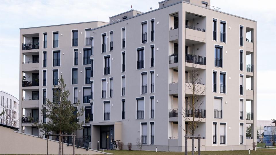 Das Bild zeigt ein modernes, mehrstöckiges Wohngebäude mit grauer Putzfassade, schmalen Fenstern mit Rollläden und französischen Balkonen sowie seitlichen Loggien.