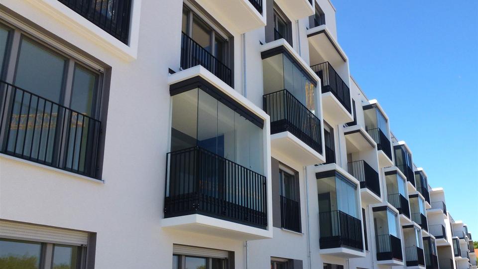 Das Bild zeigt eine moderne Hausfassade mit verglasten Erkern und schwarzen Geländern vor weißen Wänden unter klarem, blauem Himmel.